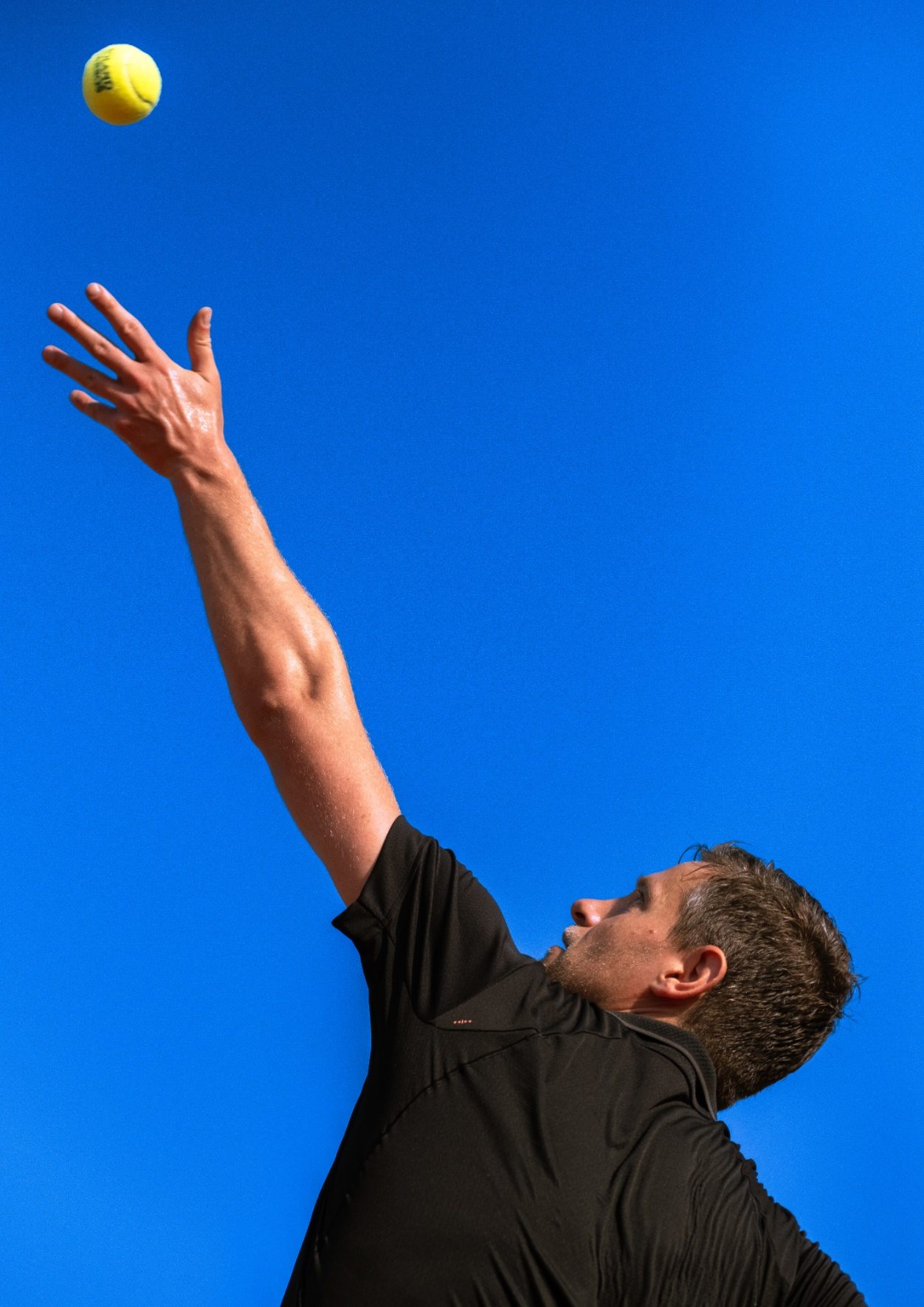Tennisser serveert een bal onder een heldere blauwe lucht. vanderstarre fotografie